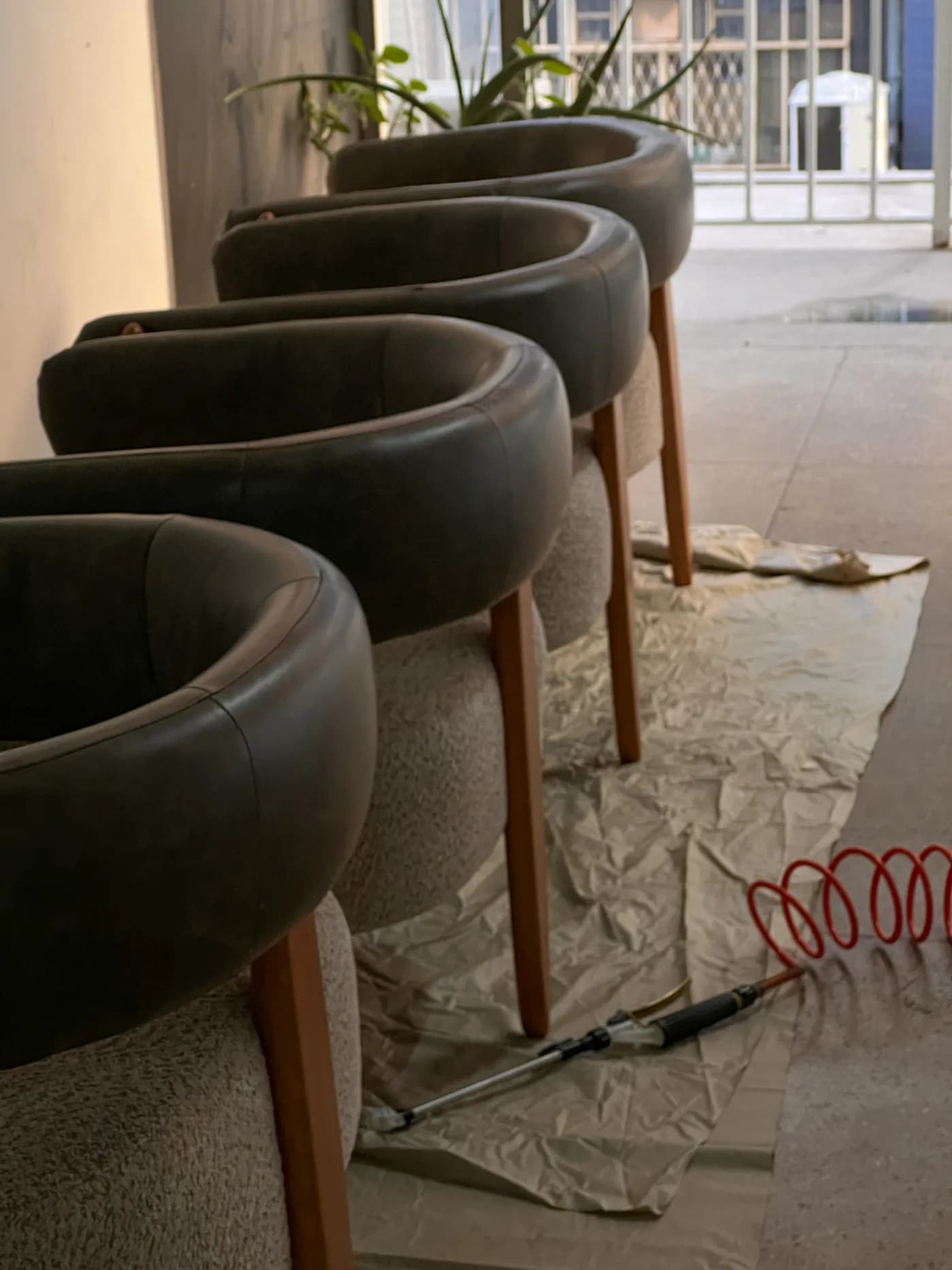 Fileira de poltronas de couro escuro com assento em bouclé sobre plástico de proteção, com a mangueira vermelha e a lança de aplicação no chão
