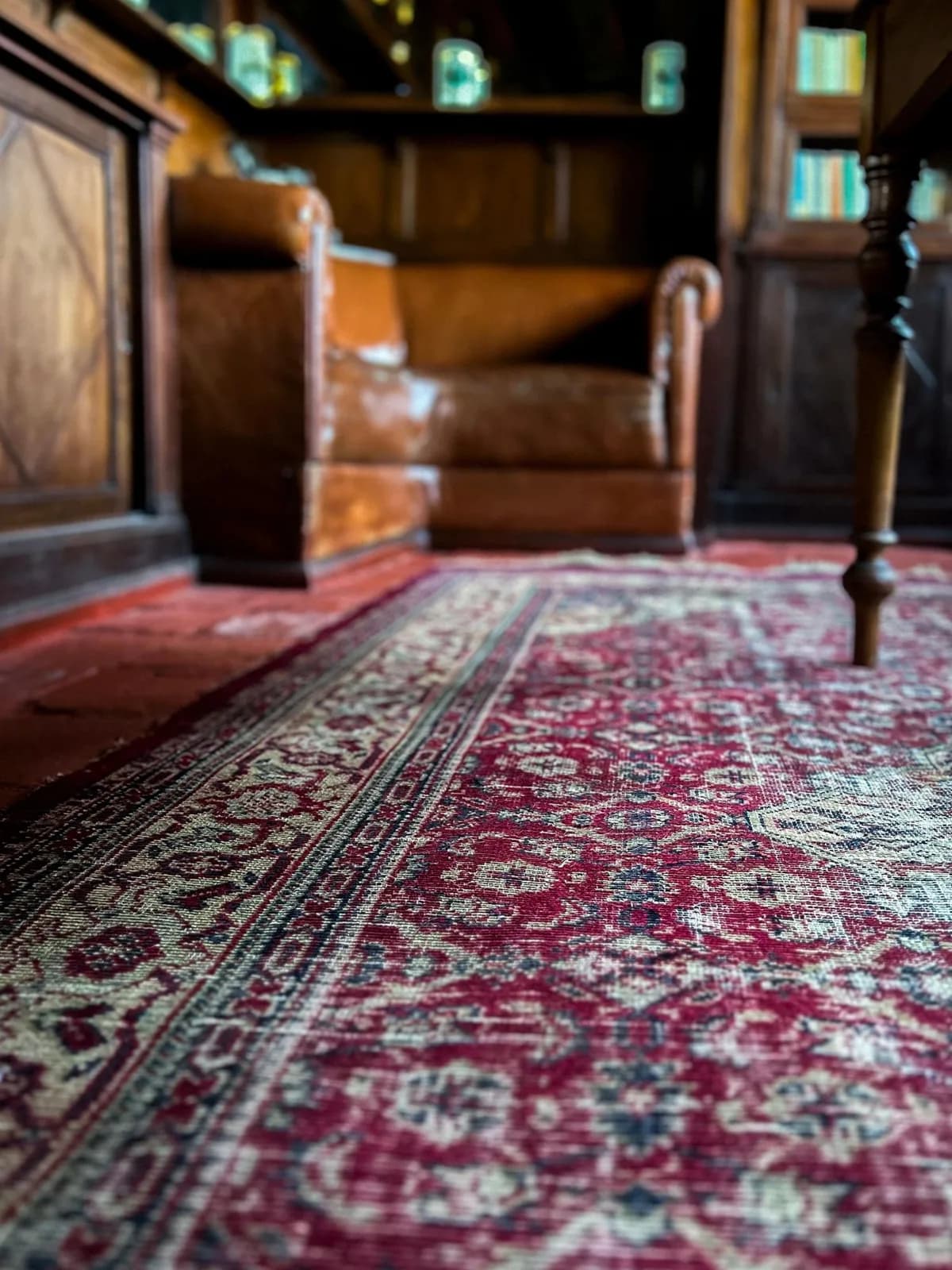 Tapete persa vermelho com sinais de desgaste, em uma sala com estantes de madeira e uma poltrona de couro ao fundo